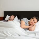 A woman lies awake next to a snoring partner in bed, illustrating the need for effective anti-snoring devices.