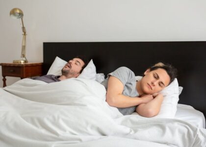 A woman lies awake next to a snoring partner in bed, illustrating the need for effective anti-snoring devices.