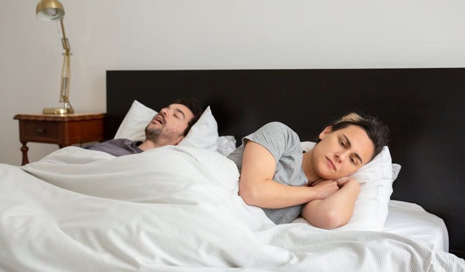 A woman lies awake next to a snoring partner in bed, illustrating the need for effective anti-snoring devices.