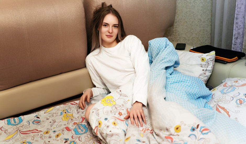 Young woman in a white shirt sits on a bed covered with patterned and blue blankets, illustrating heated blanket safety concerns.