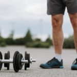 Crop anonymous male in sportswear standing near heavy metal dumbbells on asphalt road