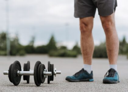 Crop anonymous male in sportswear standing near heavy metal dumbbells on asphalt road