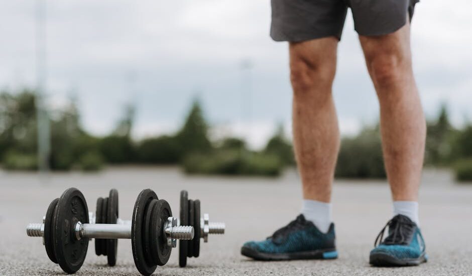 Crop anonymous male in sportswear standing near heavy metal dumbbells on asphalt road