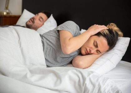 A woman looks frustrated in bed as her partner snores loudly, highlighting sleep disturbance issues.
