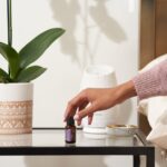 A hand reaches for a bottle of essential oil beside a white aromatherapy diffuser on a bedside table, illustrating sleep support.