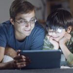 A man and a child wearing blue light glasses look at a tablet together, illustrating screen use and eye protection for the best blue light glasses for screens i