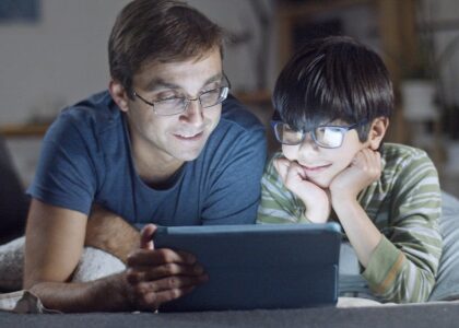 A father and son lying in bed, happily watching a tablet indoors at night.