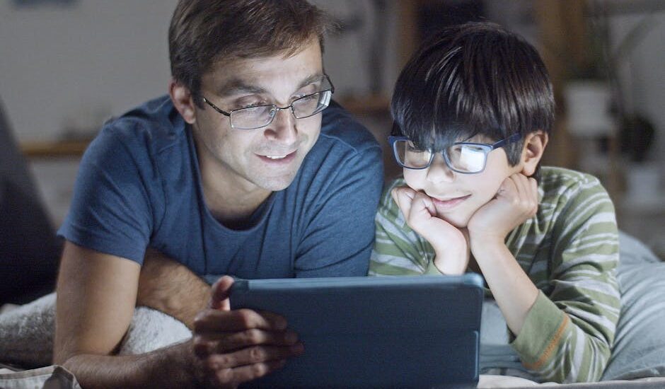 A father and son lying in bed, happily watching a tablet indoors at night.