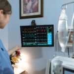 A nurse in blue scrubs examines a medical monitor displaying vital signs in a hospital setting.