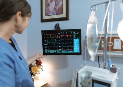 A nurse in blue scrubs examines a medical monitor displaying vital signs in a hospital setting.