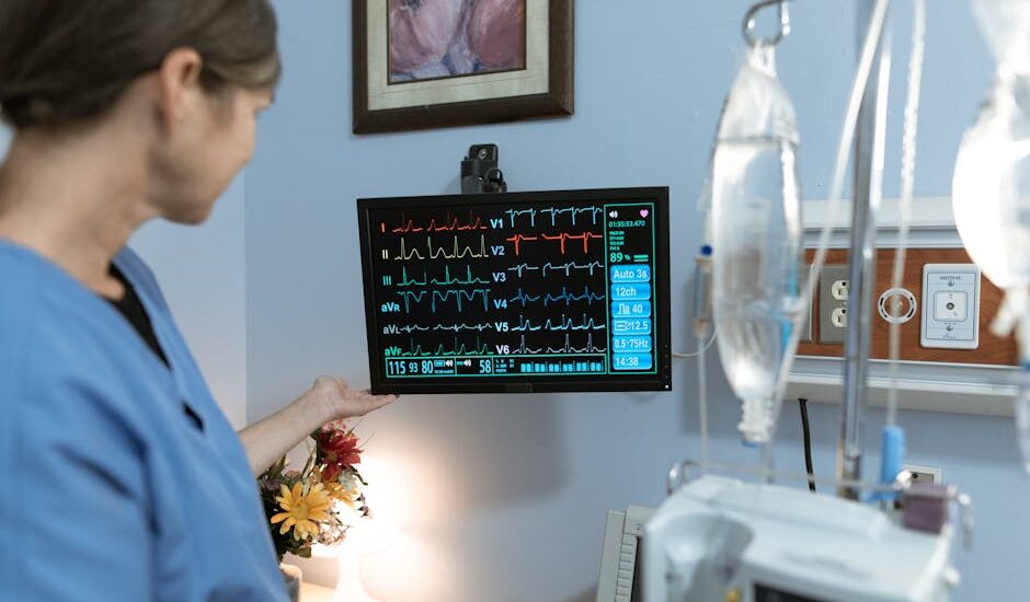 A nurse in blue scrubs examines a medical monitor displaying vital signs in a hospital setting.