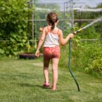 Back view of a young girl watering a garden with a hose on a sunny day.