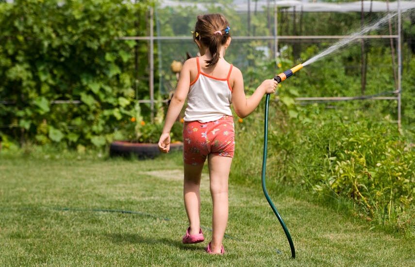 Back view of a young girl watering a garden with a hose on a sunny day.