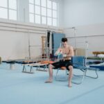 Shirtless young male gymnast preparing grips in an indoor training gym.