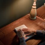 A senior woman reads a book on a wooden desk under warm lamp lighting.