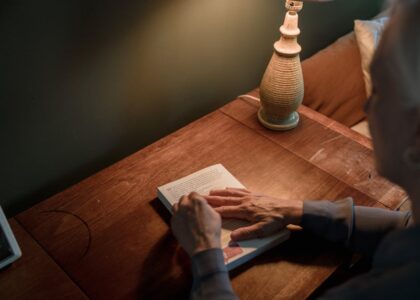A senior woman reads a book on a wooden desk under warm lamp lighting.