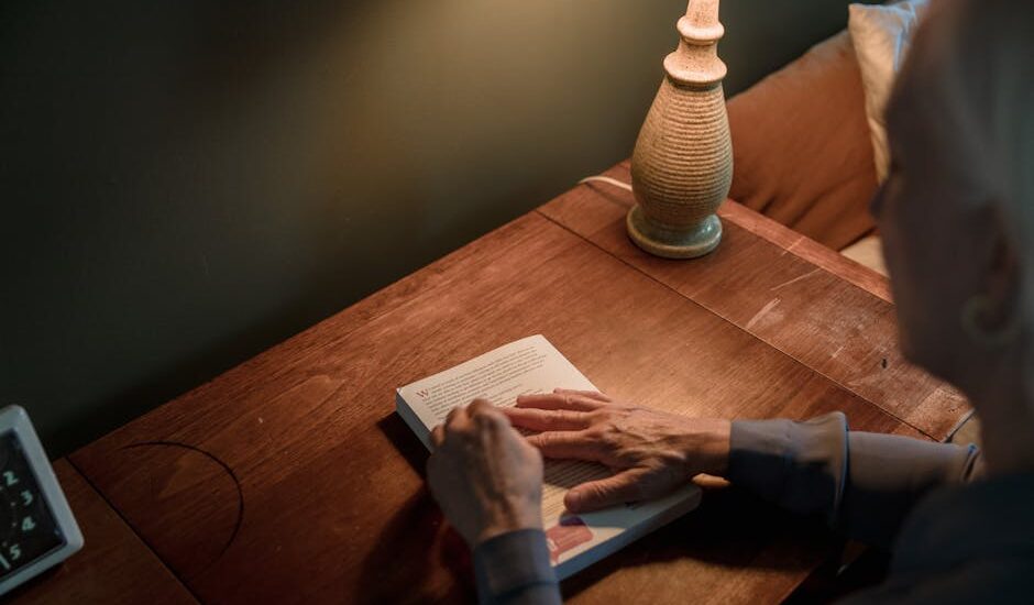 A senior woman reads a book on a wooden desk under warm lamp lighting.