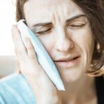 A woman in discomfort applying a cold compress to her cheek for relief from toothache pain.