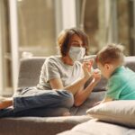 A mother wearing a face mask playing with her son on the sofa in a bright living room.