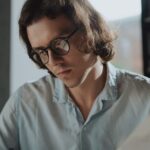 A man in a light blue shirt wears stylish reading glasses while reading a book, illustrating options for men’s reading eyewear in 2026.