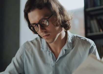 A man in a light blue shirt wears stylish reading glasses while reading a book, illustrating options for men’s reading eyewear in 2026.