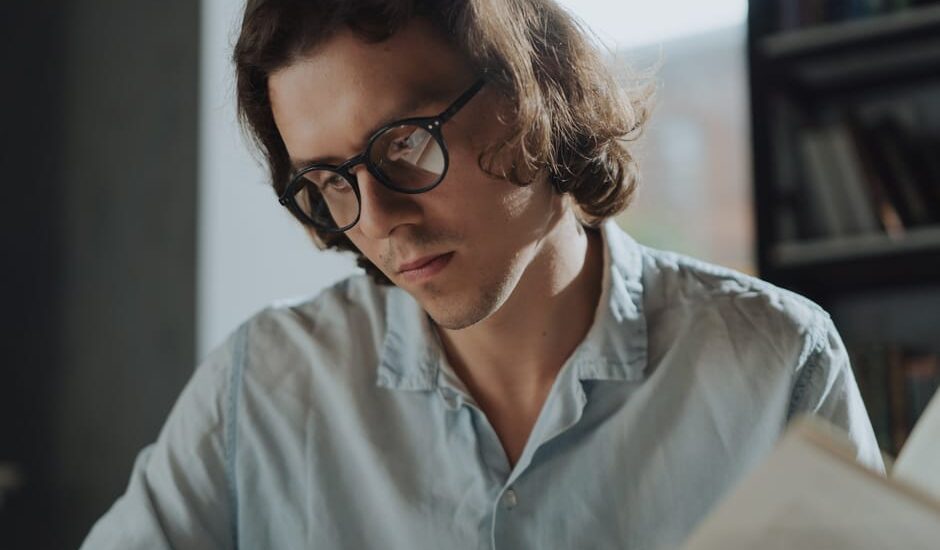 A man in a light blue shirt wears stylish reading glasses while reading a book, illustrating options for men’s reading eyewear in 2026.