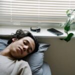A young man sleeps on a bed near a window, with a fitness tracker and smartphone on the sill, illustrating sleep tracking devices for 2026.