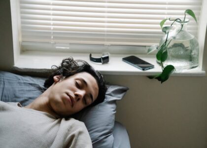 A young man sleeps on a bed near a window, with a fitness tracker and smartphone on the sill, illustrating sleep tracking devices for 2026.