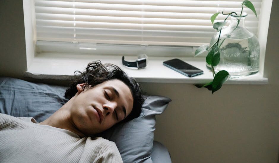 A young man sleeps on a bed near a window, with a fitness tracker and smartphone on the sill, illustrating sleep tracking devices for 2026.