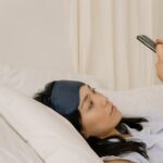 A young woman lying in bed using a smartphone with a sleep mask.