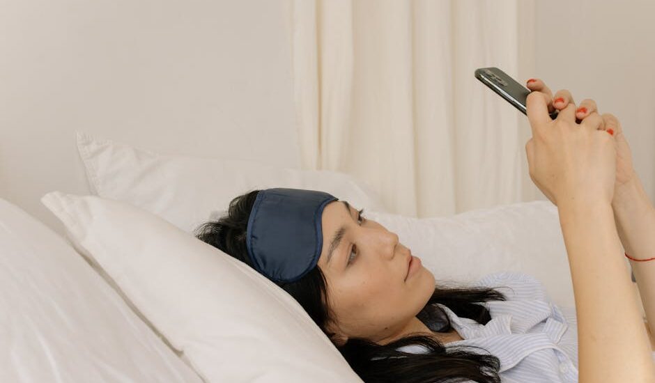 A young woman lying in bed using a smartphone with a sleep mask.