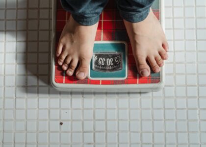 From above crop anonymous barefoot child in jeans standing on weigh scales on tiled floor of bathroom