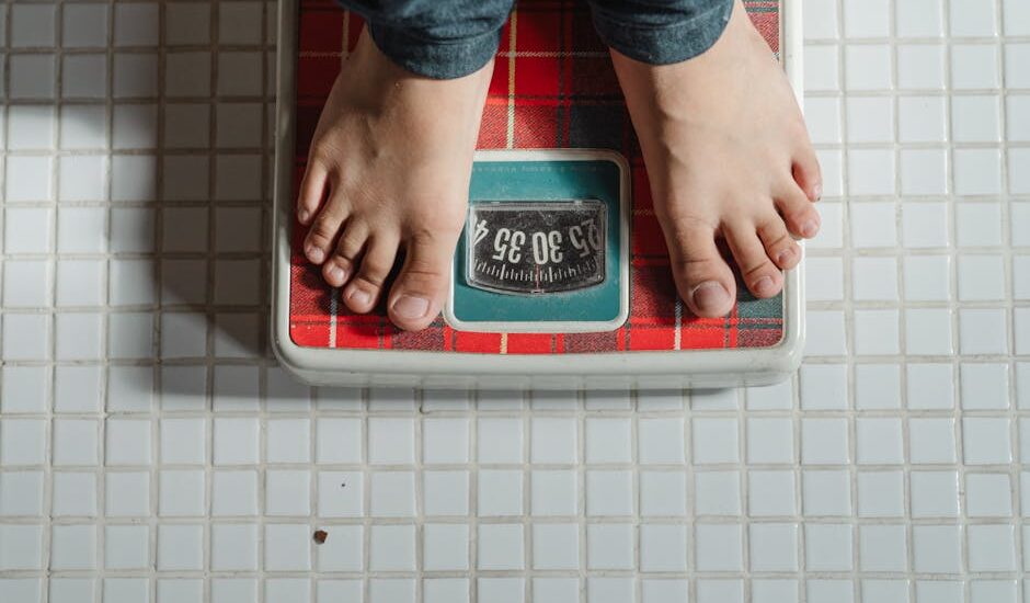 From above crop anonymous barefoot child in jeans standing on weigh scales on tiled floor of bathroom