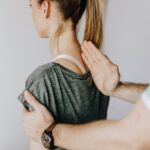 Back view of crop unrecognizable osteopath in uniform and wristwatch checking up back of slim female patient in casual wear on white background