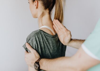 Back view of crop unrecognizable osteopath in uniform and wristwatch checking up back of slim female patient in casual wear on white background