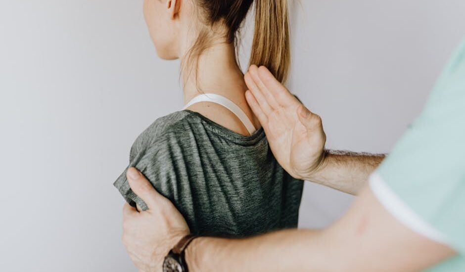 Back view of crop unrecognizable osteopath in uniform and wristwatch checking up back of slim female patient in casual wear on white background
