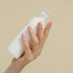 A close-up of a hand holding a white cosmetic bottle against a neutral background, perfect for product mockups and beauty industry use.