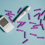 Cholesterol test kit components, including a digital meter, lancing device, and purple lancets, arranged on a blue background for a review article.
