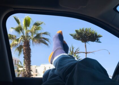 Person wearing blue and orange compression socks with feet up in a car, highlighting comfort for travel against a sunny palm tree view.
