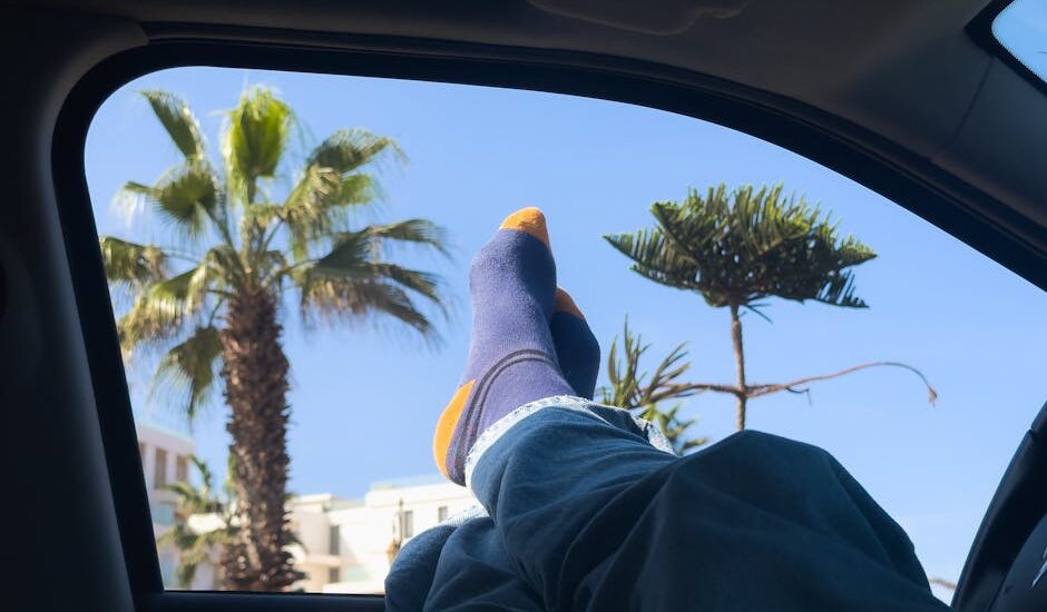 Person wearing blue and orange compression socks with feet up in a car, highlighting comfort for travel against a sunny palm tree view.