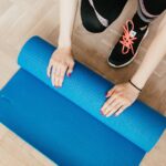 From above of unrecognizable female athlete in leggings and sneakers unfolding sport mat for practicing yoga in morning at home
