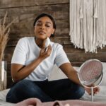 A woman sits on a bed using an electric facial massager, illustrating self-care practices related to electric eye massager benefits.