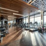 Modern exercise bikes and treadmills and various fitness equipment in spacious gym with wooden floor and panoramic windows in sunlight