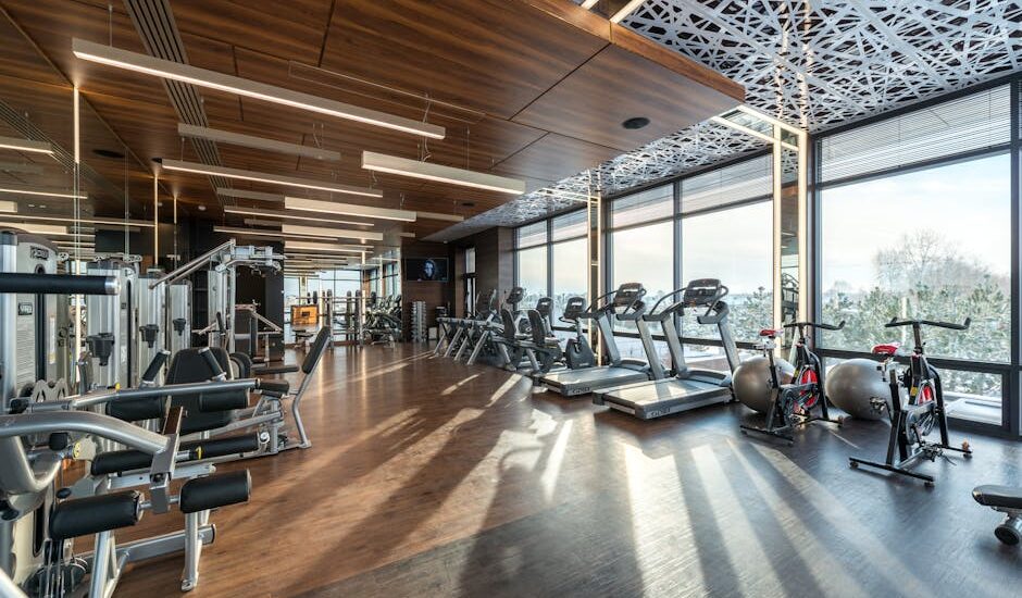 Modern exercise bikes and treadmills and various fitness equipment in spacious gym with wooden floor and panoramic windows in sunlight