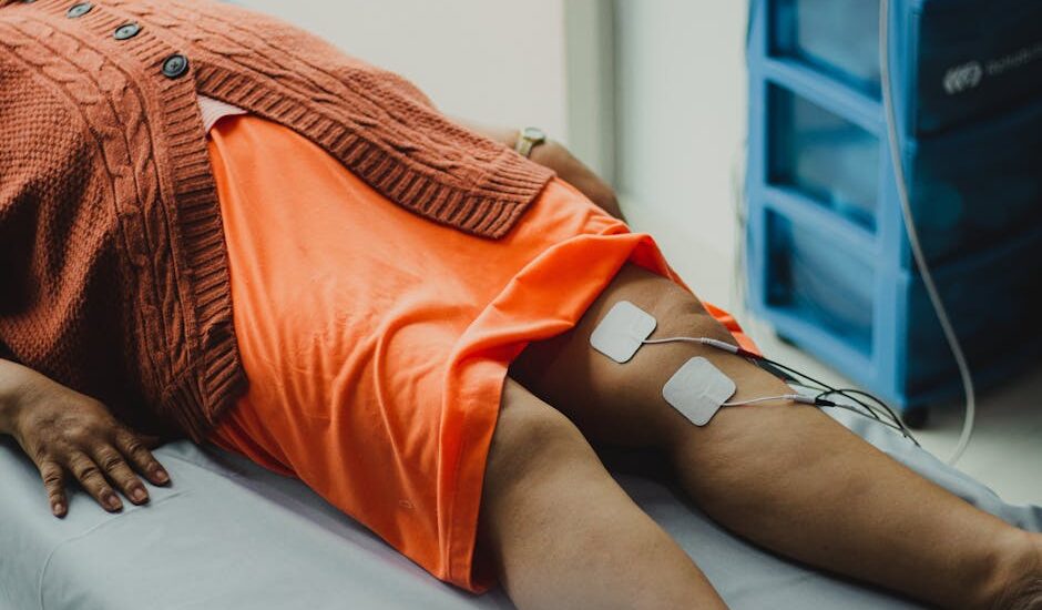 Person lying on a medical table with electrode pads attached to their thighs, illustrating EMS and TENS pain relief methods.