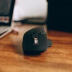 Close-up of a sleek, black wireless computer mouse on a wooden desk, next to a laptop.
