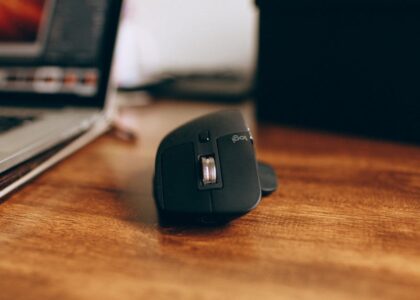 Close-up of a sleek, black wireless computer mouse on a wooden desk, next to a laptop.