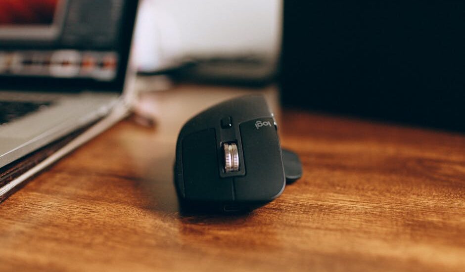 Close-up of a sleek, black wireless computer mouse on a wooden desk, next to a laptop.