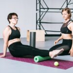 Two women in athletic wear demonstrate foam roller and recovery tool use, illustrating options for muscle relief discussed in the foam roller vs massage stick a