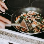 A person stirring vibrant vegetables in a frying pan on an electric stove.